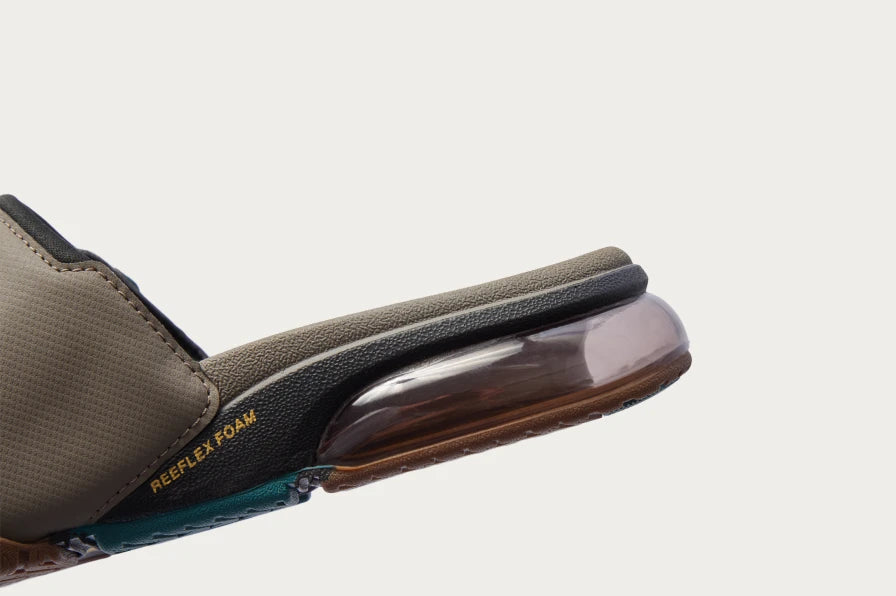Side view of brown and black slide sandal facing right, featuring transparent cushioned heel and textured REEF footbed.